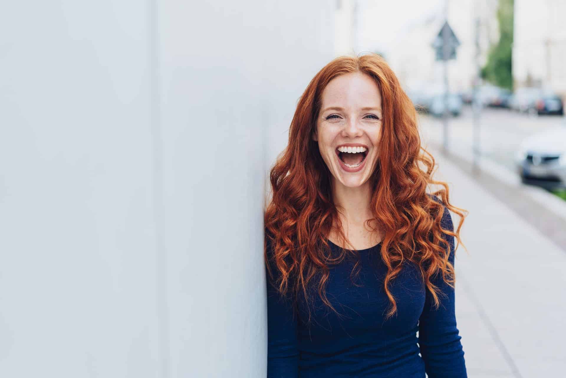 Eine rothaarige junge Frau lehnt sich an eine Wand und lacht. Dabei zeigt sie ihr schönes harmonisches Lächeln.